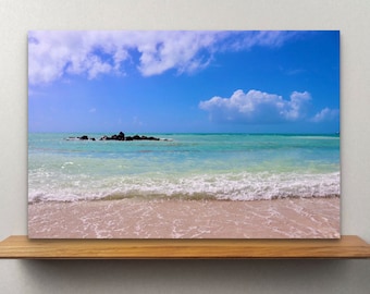 May include: A scenic canvas print of a tropical beach scene. The image features a bright blue sky with fluffy white clouds, turquoise water, and a sandy beach. A small cluster of rocks is visible in the distance.