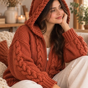 Peut inclure: Un pull à capuche zippé de couleur rouille à manches longues, doté d'un motif en tricot torsadé proéminent sur les manches et la capuche, avec des poignets côtelés et une fermeture éclair métallique allant du col à l'ourlet.
