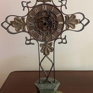 May include: Ornate, cross-shaped clock with a bronze-colored face and Roman numerals. The clock has a decorative metal frame with leaf-like accents and stands on a detailed base. The clock's hands indicate the time.