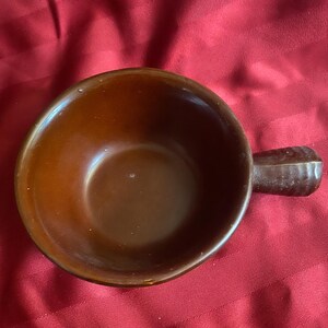 May include: A brown ceramic soup bowl with a handle. The bowl has a rounded shape and a smooth, glossy finish. The handle is rectangular and attached to the side of the bowl. The bowl is set against a red background.