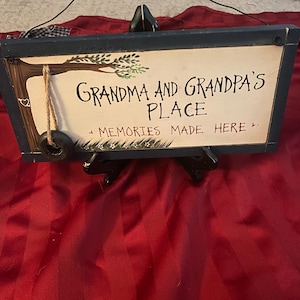 May include: Wooden sign with the text "Grandma and Grandpa's Place" and "Memories Made Here." The sign has a dark blue frame, a tree illustration, and a rope swing detail. The background is a red fabric.