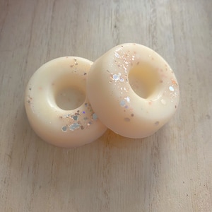 Peut inclure: Deux fondants de cire en forme de beignets blancs avec une finition scintillante. Les fondants sont sur une surface en bois clair.