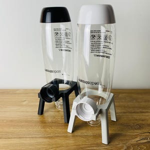 Könnte beinhalten: Zwei durchsichtige SodaStream-Flaschen aus Kunststoff mit schwarzen und weißen Sockeln. Die Flaschen sind mit "SodaStream" beschriftet und tragen gedruckte Sicherheitshinweise.