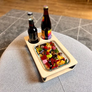 May include: A wooden serving tray with two bottle holders and a rectangular glass dish filled with colorful gummy candies. Two dark bottles are in the holders. The tray sits on a gray upholstered ottoman.