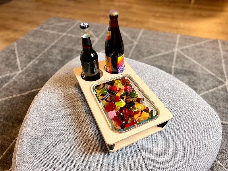May include: A wooden serving tray with two bottle holders and a rectangular glass dish filled with colorful gummy candies. Two dark bottles are in the holders. The tray sits on a gray upholstered ottoman.