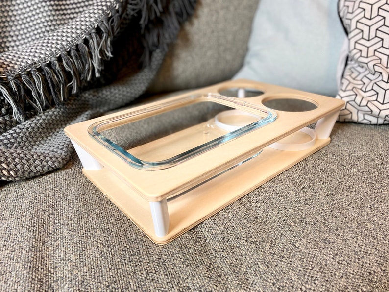 May include: A light-colored wooden serving tray with a rectangular glass dish and two circular cutouts. The tray has a lower shelf with two white supports and is designed to hold drinks and snacks. The tray is on a gray surface.