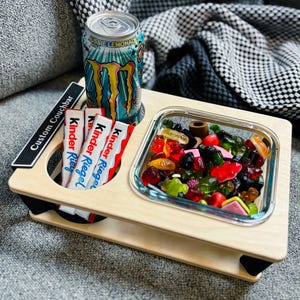 Könnte beinhalten: Ein Couchtisch aus Holz mit Getränkehalter, Süßigkeitenfach und einer Glasschale voller bunter Gummibonbons. Der Tisch hält eine Dose Aussie Lemonade und drei Kinder Riegel Schokoriegel. Der Text "Custom Couchbar" ist auf einem schwarzen Etikett aufgedruckt.