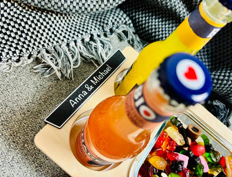 May include: A wooden serving tray with two bottles of soda and a small dish of colorful candies. The bottles are orange and yellow, with blue and white bottle caps. The tray has the text "Anna & Michael".