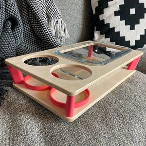 May include: A wooden serving tray with a natural finish and bright pink accents. The tray has cutouts for two glasses and a rectangular dish. The tray is supported by pink legs and is designed for serving food and drinks.