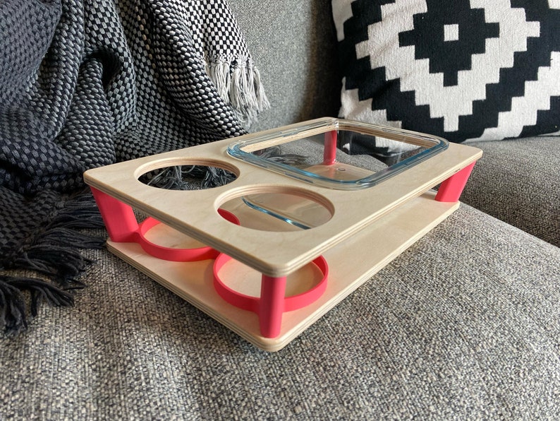 May include: A wooden serving tray with a natural finish and bright pink accents. The tray has cutouts for two glasses and a rectangular dish. The tray is supported by pink legs and is designed for serving food and drinks.