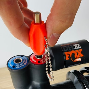 Könnte beinhalten: Nahaufnahme eines Fox DHx2 Factory Series Dämpfers mit einem roten und blauen Einstellknopf. Ein kleines orangefarbenes Werkzeug mit einem Schlüsselanhänger ist am Dämpfer befestigt.