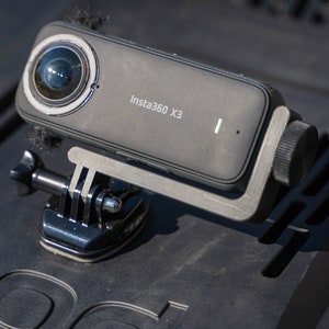 May include: A black Insta360 X3 action camera mounted on a black GoPro-style mount. The camera has a large lens on the front and a small screen on the side.
