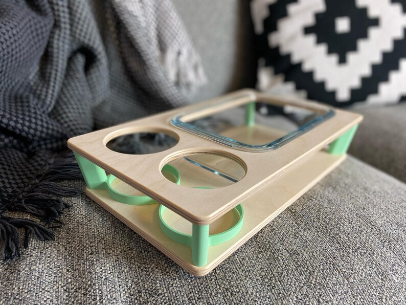 May include: A wooden serving tray with a natural finish and mint green accents. The tray features a rectangular glass dish, two circular cutouts, and two recessed areas for holding items. The tray is designed for convenience and style.