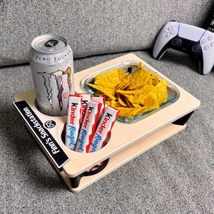 Könnte beinhalten: Ein hölzernes Snacktablett mit einer Dose Monster Energy Drink, einer Glasschale mit Tortilla-Chips und mehreren Kinder Riegel-Riegeln. Im Hintergrund ist ein weiß-schwarzer Gamecontroller zu sehen. Auf dem Tablett steht "Finn's Snackstation".