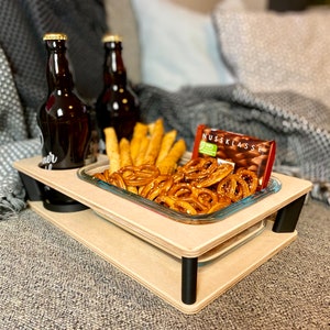 May include: A two-tiered wooden serving tray with black legs, holding two brown bottles, a glass dish of pretzels and breadsticks, and a chocolate bar. The tray is on a gray couch.