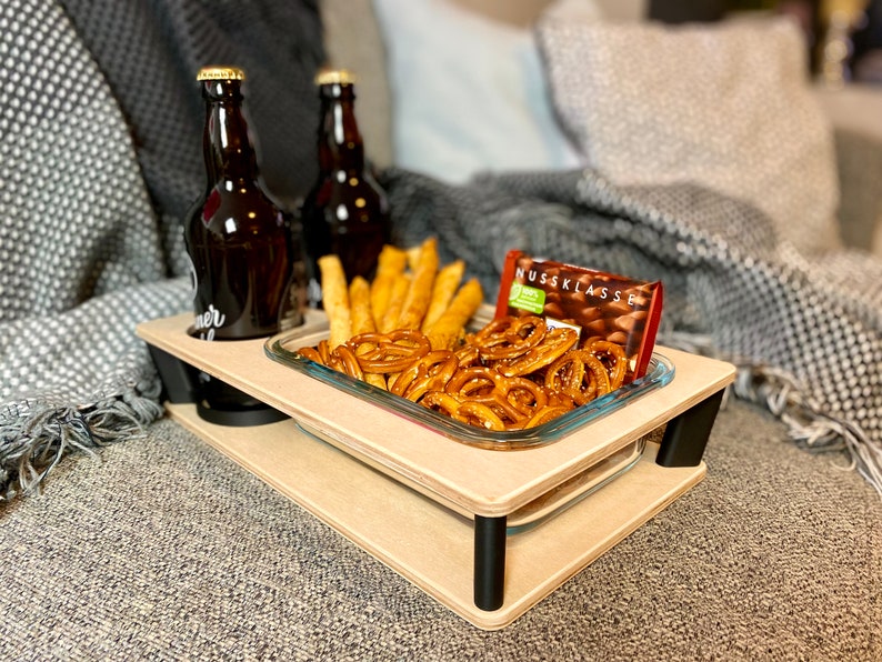 May include: A two-tiered wooden serving tray with black legs, holding two brown bottles, a glass dish of pretzels and breadsticks, and a chocolate bar. The tray is on a gray couch.