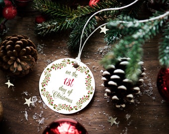 Może przedstawiać: Okrągła biała ozdoba z zielonym wieńcem i czerwonymi jagodami wokół krawędzi. Ozdoba zawiera napis "on the 1st day of Christmas" czerwonymi i czarnymi literami.