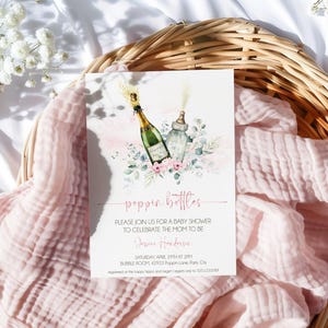 May include: A pink and white baby shower invitation featuring a watercolour illustration of a champagne bottle and a baby bottle. The text on the invitation reads "poppin' bottles" and includes the date, time, and location of the event.