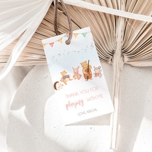 Puede incluir: Una etiqueta de regalo con una ilustración de acuarela de animales del bosque tocando instrumentos musicales. La etiqueta tiene un fondo azul claro con notas musicales y banderines. El texto dice "THANK YOU FOR playing WITH ME" y "LOVE, ABIGAIL."