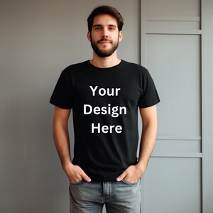 May include: A man wearing a black t-shirt with the text "Your Design Here" printed in white on the front. He is standing in front of a gray wall with a white door.