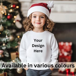 May include: A white sweatshirt with the text "Your Design Here" printed on the front. The sweatshirt is being modeled by a child wearing a red Santa hat. The child is standing in front of a Christmas tree.