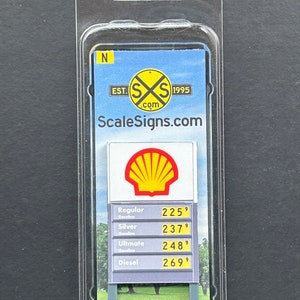 Puede incluir: Un letrero de tren modelo para una gasolinera con un logotipo de Shell y precios para gasolina regular, plata, ultimate y diésel. El letrero es gris con texto amarillo y fondo blanco. El letrero está en un paquete de plástico con el nombre de la empresa "ScaleSigns.com" impreso en él.