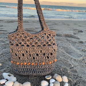 Puede incluir: Una bolsa de mano de crochet gris con cuentas marrones alrededor de la parte superior. La bolsa está sentada en una playa de arena con conchas esparcidas a su alrededor.