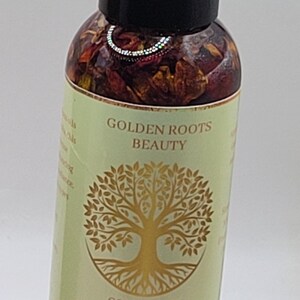 May include: A clear glass bottle of Golden Roots Beauty Golden Glow Elixir. The bottle has a black cap and is filled with reddish-brown botanicals. The label features a gold tree design and text that reads "Nourish, Hydrate and Illuminate Your Skin". The bottle contains 120 ml.