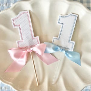 May include: Two number one cake toppers featuring embroidered fabric numbers. The left topper has a pink stitched border and a pink satin bow on a wooden stick. The right topper has a light blue stitched border and a light blue satin bow on a clear stick.