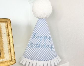 May include: A conical party hat featuring a light blue and white gingham pattern with the embroidered text Happy Birthday in blue thread. The hat is topped with a large white pom-pom and finished with a white pleated ribbon trim at the base.