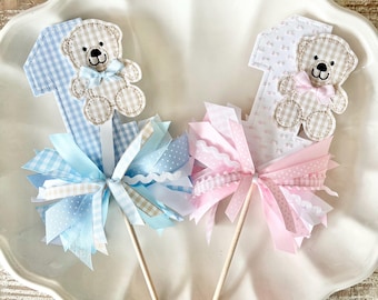 May include: Two decorative number one cake toppers. One is blue gingham with a teddy bear and blue bow, the other is white with a teddy bear and pink bow. Both have ribbon accents in coordinating colors.