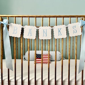 May include: A white baby crib with a gold metal frame. A blue and white banner with the name "BANKS" is hanging above the crib. The crib is decorated with a blue and white gingham pillow and a red, white, and blue American flag.