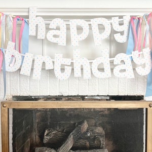 May include: A white paper banner with the words "happy birthday" in white letters with blue polka dots. The banner is decorated with colorful ribbons.