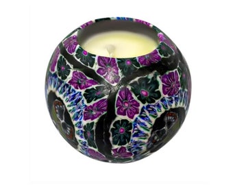 May include: A round candle with a black, white, purple, and green floral design. The candle is lit and the wax is melting.