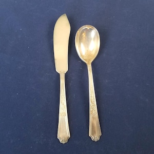 Op de afbeelding: Zilveren bestekset met een botermes en een lepel. Het botermes heeft een gebogen lemmet en een decoratief handvat. De lepel heeft een ovale kom en een bijpassend decoratief handvat. Beide stukken zijn zilverkleurig.