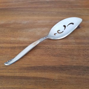 May include: A silver serving spoon with a slotted, oval head and a long, slender handle. The spoon features decorative detailing on the handle and three curved cutouts on the head. The spoon is designed for serving food.