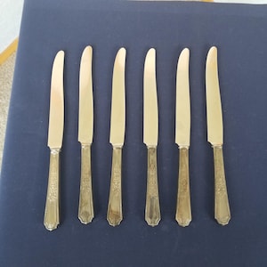 Puede incluir: Juego de seis cuchillos plateados con un patrón floral decorativo en los mangos. Las hojas son lisas y curvas. Los cuchillos están colocados sobre una superficie azul oscuro, lo que sugiere un ambiente de cena formal.