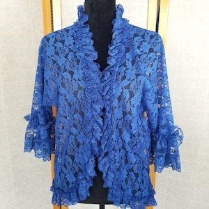 Könnte beinhalten: Eine leuchtend blaue Spitzenjacke mit gerüschtem Kragen und Manschetten. Die Jacke hat ein Blumenmuster und ein offenes Design. Die Ärmel sind dreiviertellang, und die Jacke wird auf einer schwarzen Schaufensterpuppe präsentiert.