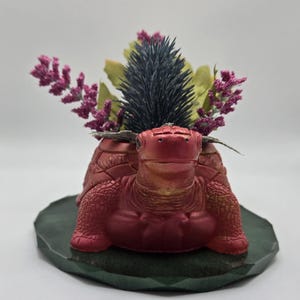 May include: A red turtle-shaped planter with a metallic finish, holding a floral arrangement. The arrangement includes purple, green, and blue-grey dried flowers. The planter sits on a dark green, geometric-edged base.