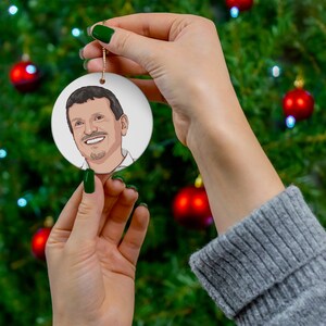 May include: A white round Christmas ornament with a cartoon drawing of a man with brown hair and a beard. The ornament is hanging from a gold string.