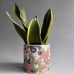 May include: A cylindrical white ceramic planter featuring a colourful botanical pattern of pink, dark grey, and olive green flowers and leaves, containing a tall snake plant with dark green sword-shaped leaves and bright yellow edges.