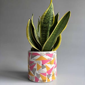 May include: A cylindrical ceramic planter featuring a white base with a repeating geometric pattern of pink triangles, yellow triangles, and small black stippled dots. A tall snake plant with dark green leaves and yellow edges grows from the pot.