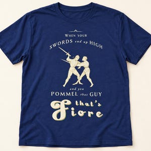 May include: A navy blue short-sleeve t-shirt featuring a cream-coloured graphic of two figures fencing with swords. The text reads: WHEN YOUR SWORDS end up HIGH and you POMMEL that GUY that's Fiore. The design is centred on the front of the shirt.