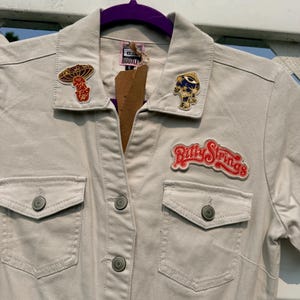 May include: A beige button-down shirt with two chest pockets and metal buttons. The collar features a mushroom patch and a celestial eye patch. A red and white Billy Strings patch is stitched above the right chest pocket. A tag is attached.