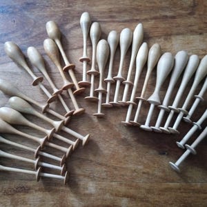May include: A collection of wooden lace bobbins, each with a rounded top and a spool-like base. The bobbins are arranged on a wooden surface, showcasing their light brown color and smooth texture. These tools are used in lacemaking.