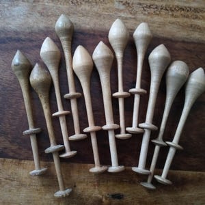 May include: A collection of wooden lace bobbins, each with a bulbous head and a slender stem. The bobbins are arranged on a dark wooden surface, showcasing their natural wood grain and unique shapes. These tools are used in lacemaking.