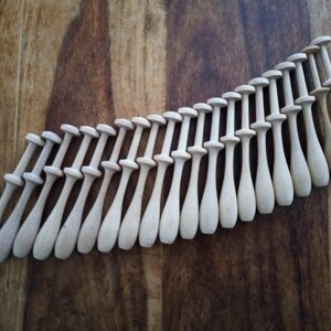 May include: A collection of twenty-one wooden lace bobbins, arranged diagonally on a dark wood surface. Each bobbin has a rounded head, a narrow neck, and a tapered body. The bobbins are made of light-colored wood.