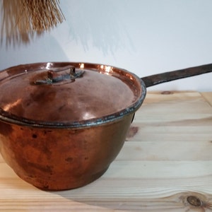 Könnte beinhalten: Ein antiker Kupfertopf mit Deckel und einem langen Eisenhenkel. Der Topf hat einen abgerundeten Boden und einen leicht ausgestellten Rand. Der Topf steht auf einer Holzoberfläche.