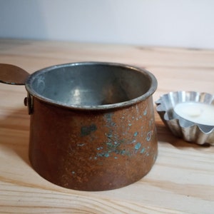 May include: A small copper pot with a handle and a lid. The pot has a patina of green and brown. It is sitting on a wooden surface next to a small silver candle holder.