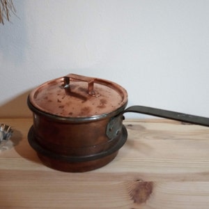 Könnte beinhalten: Ein Kupfertopf mit Deckel und langem Griff. Der Topf steht auf einer Holzoberfläche.